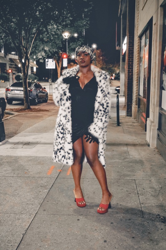 Woman in a spotted faux fur coat, black mini dress and red heels standing square to camera on a night sidewalk.