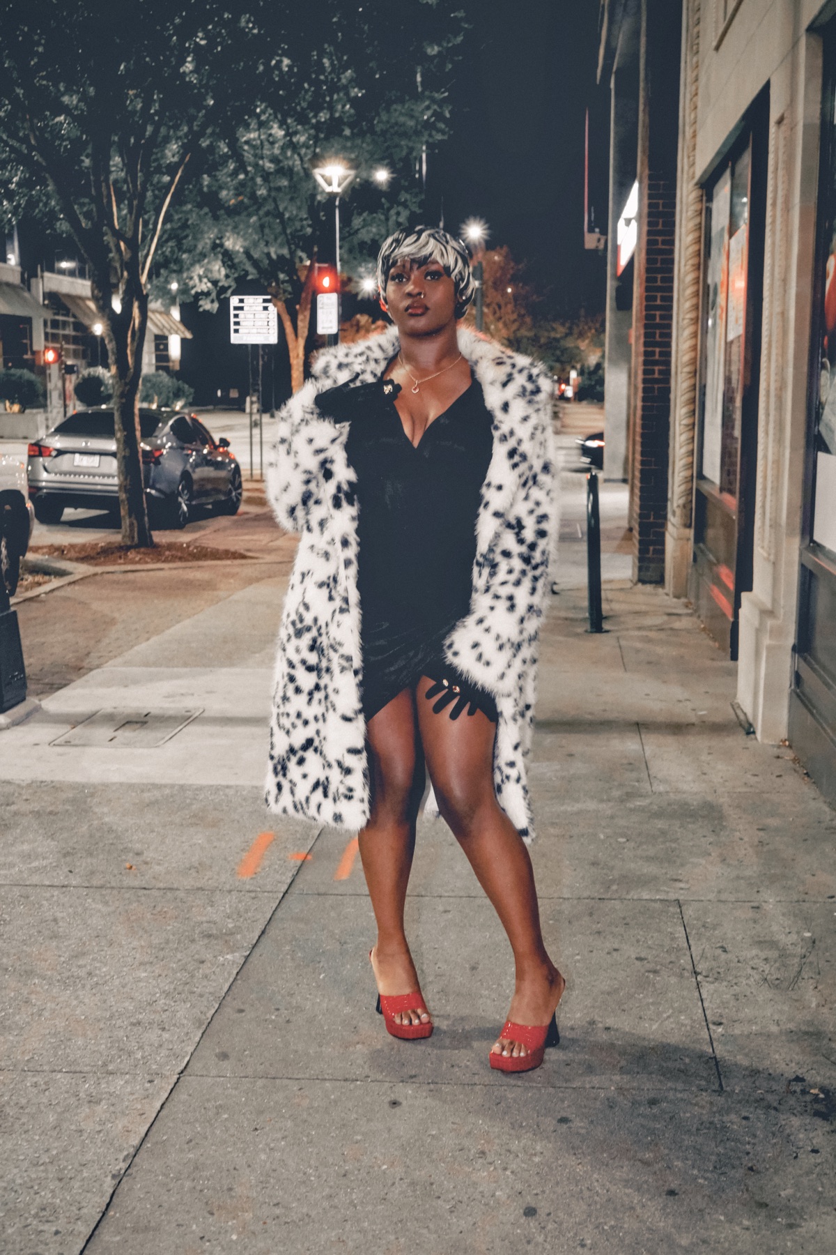 Woman in a spotted faux fur coat, black mini dress and red heels standing square to camera on a night sidewalk.