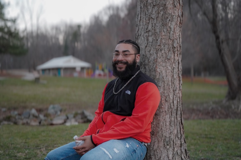 Bearded man in a red fleece leaning against a tree in a park, smiling with a chain necklace.