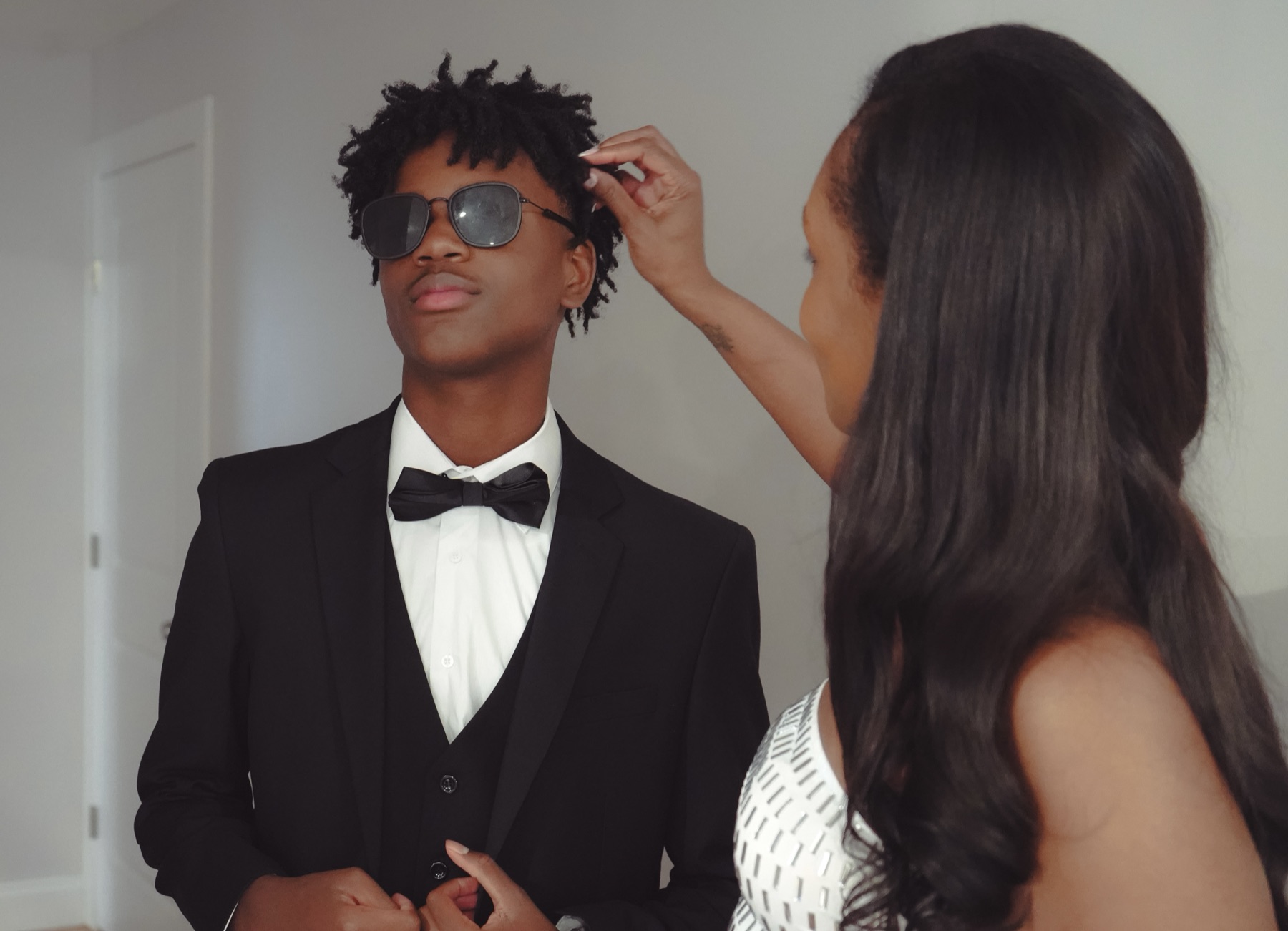 Mother in a white beaded dress adjusting her son's collar as he stands in a black tuxedo before the night out.