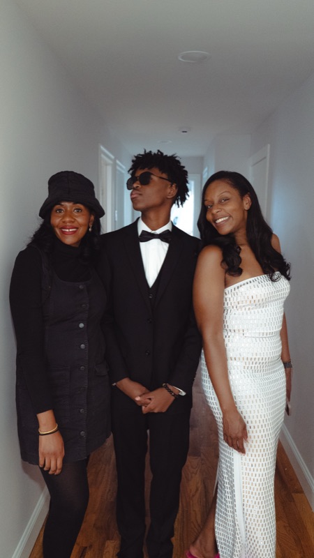 Son in a black tuxedo flanked by two women in a bright hallway before prom.