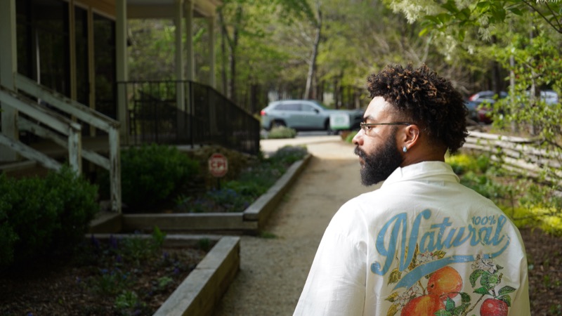 Man with glasses and beard looking over his shoulder in a white jacket on a wooded path.