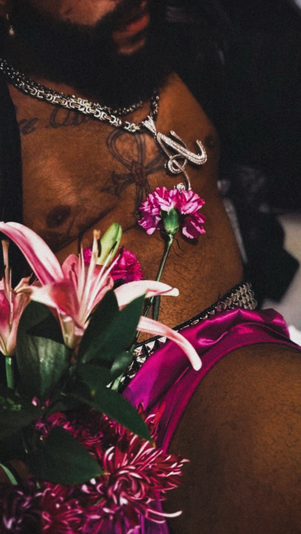 Close crop of a man's chest with silver chains and a carnation, pink lilies and magenta satin in foreground.