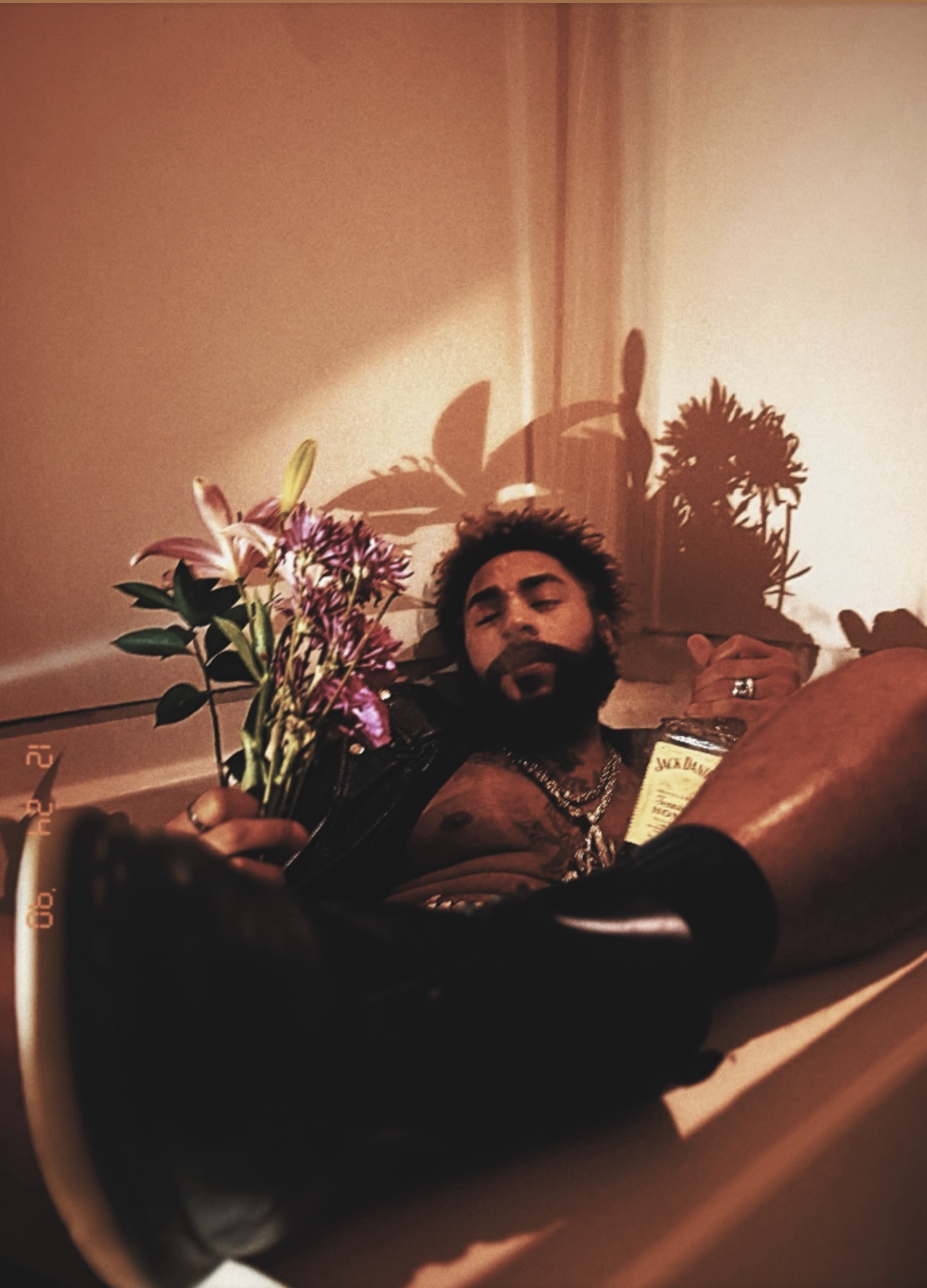 Warm-toned portrait of a bearded man reclining in a bathtub in a leather jacket, holding lilies.