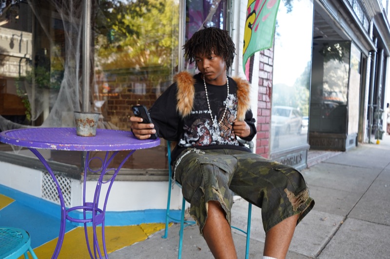 Young man in a fur-trimmed vest checking his phone at a purple café table outside a brick storefront.
