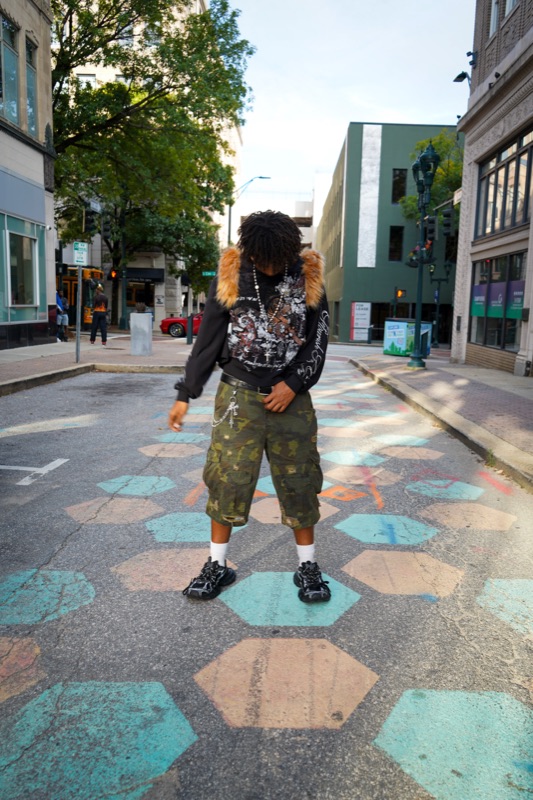 Young man in a fur-collared graphic sweatshirt and camo shorts looking down at painted hexagons on a downtown street.