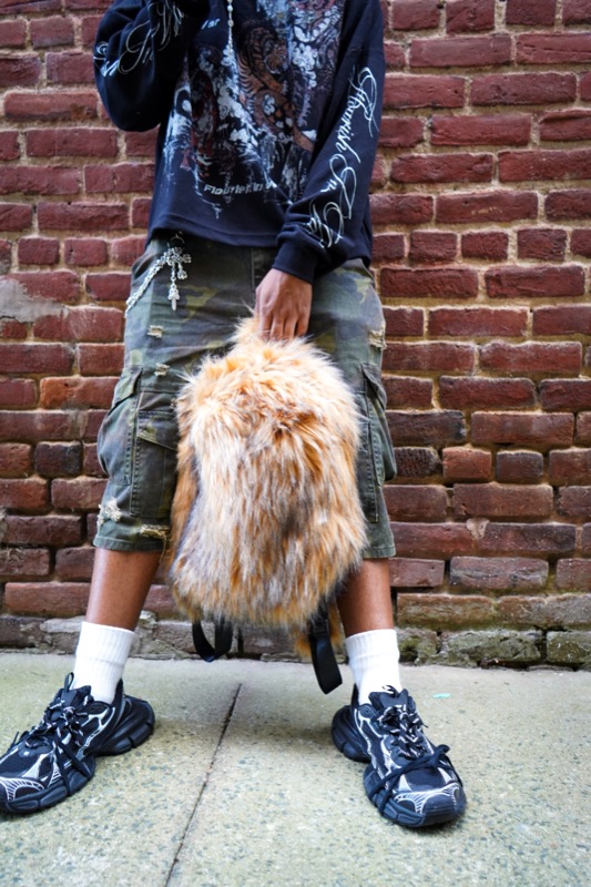 Fur backpack held against camo shorts and chunky sneakers in front of a red brick wall.