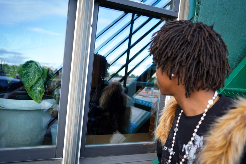 Young man with locs and a fur-trim collar studying his reflection in a green-framed shop window.