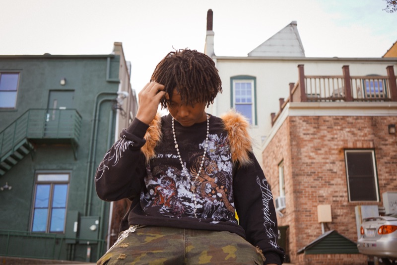 Young man with locs running a hand through his hair, head bowed, between green and brick buildings.