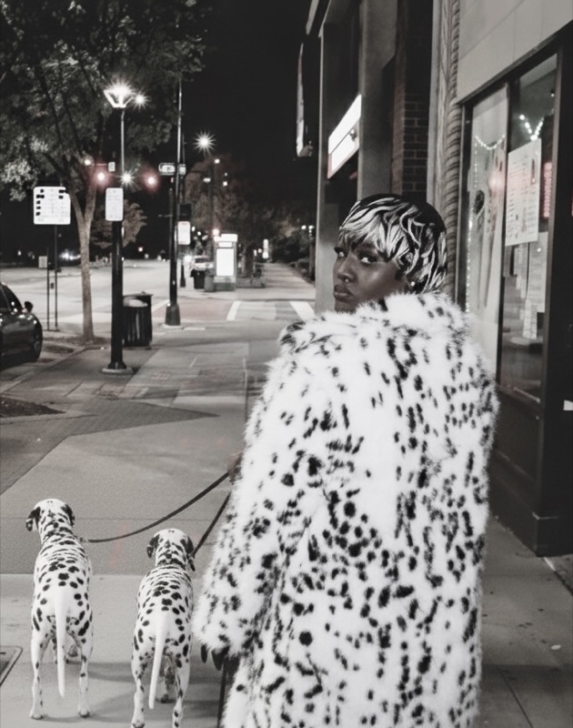 Black-and-white night scene of a woman in a dalmatian-print faux fur coat walking two dalmatians down a city sidewalk.