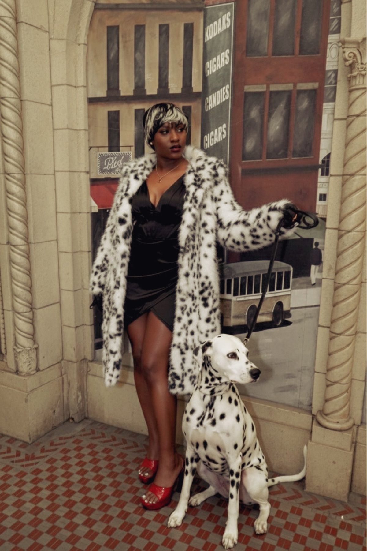 Woman in a spotted faux fur coat and red heels holding a dalmatian on a leash before a vintage painted mural.